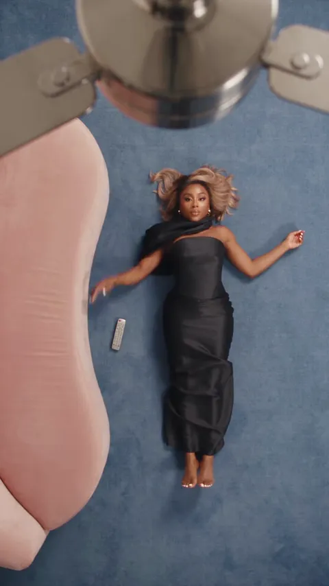 Woman in a black strapless gown lying on a blue rug beside a pink sofa, seen from the ceiling fan above.